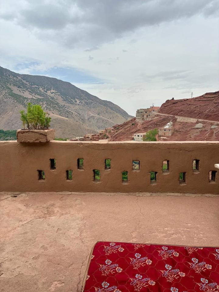 A mud-walled terrace with small window openings, a red cushion and mountains beyond