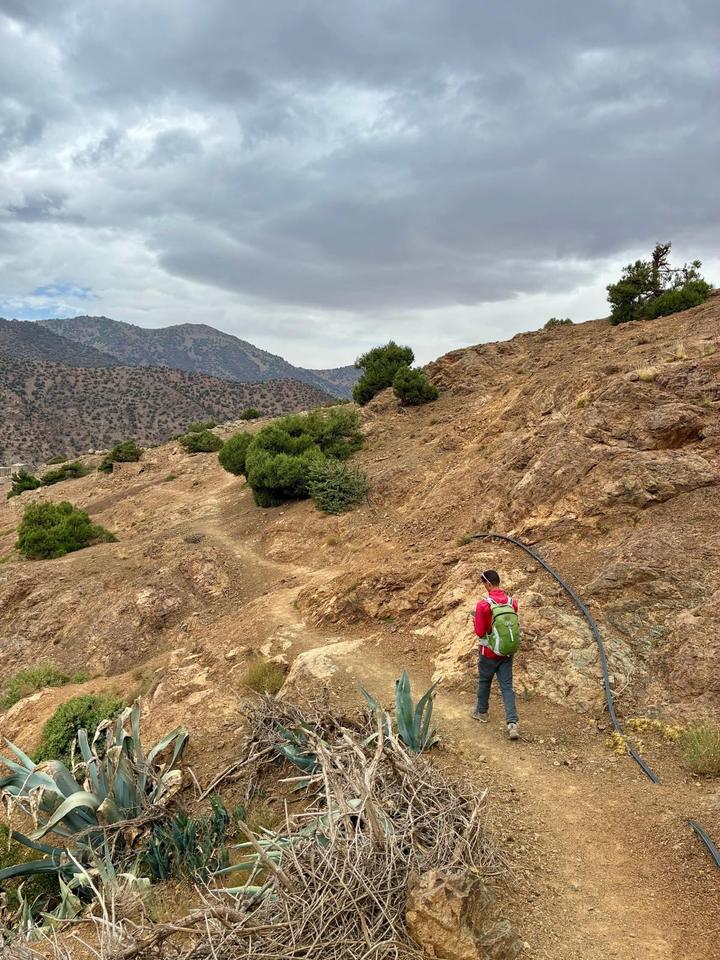 A guide in a red jacket walking a dry mountain trail under grey clouds