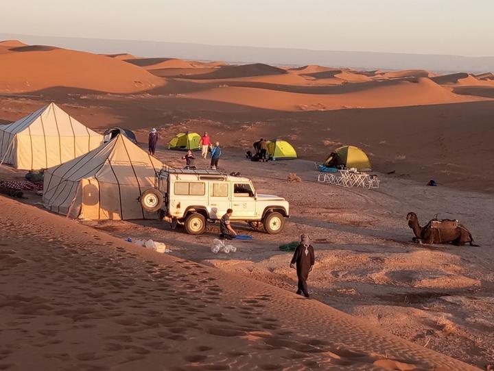 A desert camp at sunrise with tents, a four-wheel-drive vehicle and a camel