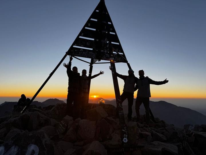 Four trekkers silhouetted against the sunrise at the summit marker of Toubkal
