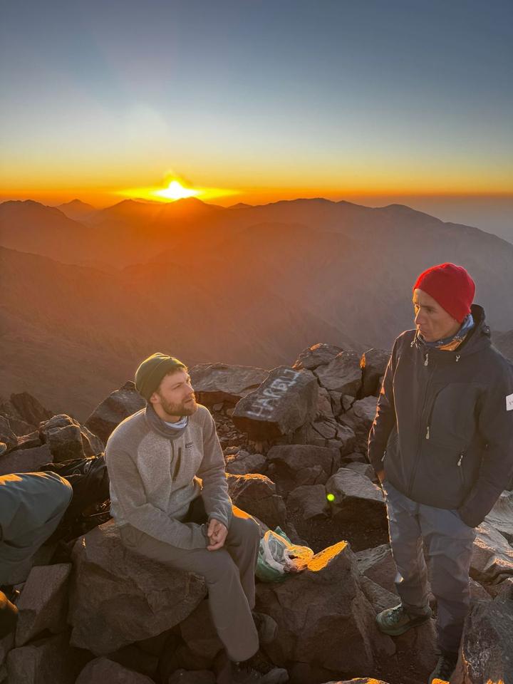 Two men talking on a high summit as the sun rises over the mountains